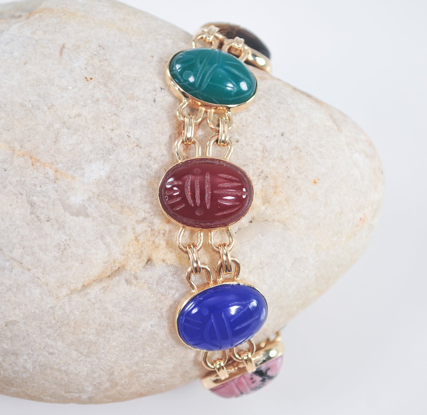 A vintage 14K gold double link scarab bracelet with a series of carved stones in various colors including red, blue, green, and yellow, displayed on a rock.